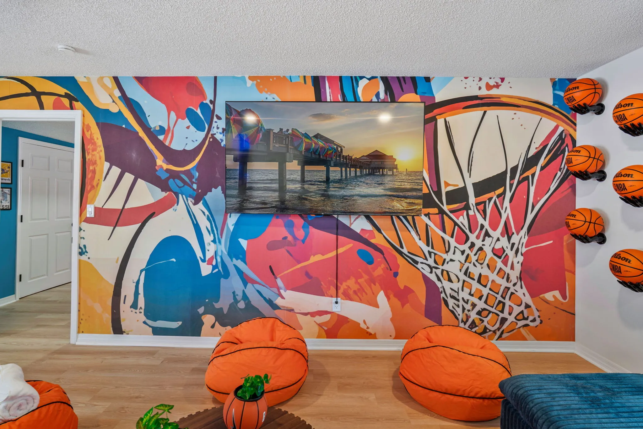 Indoor room with colorful basketball-themed wall mural, three basketball bean bags, mounted TV showing a pier sunset.